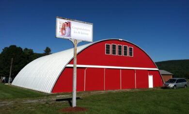 large quonset hut hangar with red endwall and sliding doors and sign in front with picture of couple and words 