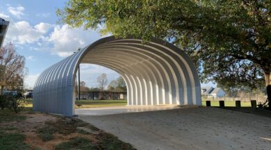 S-Model Quonset hut with open-ended walls, a concrete ramp leading into it, a black fence behind it, and a large tree to the right.