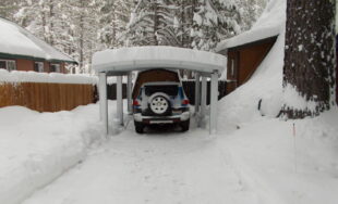A steel carport with a corrugated roof covered in snow with a blue jeep parked underneath