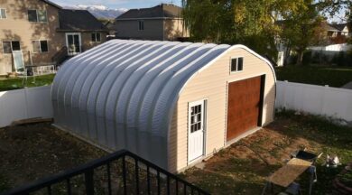 A-Model Quonset hut with tan end wall, brown garage door, white man door, and black railing going down in the front of the photo.
