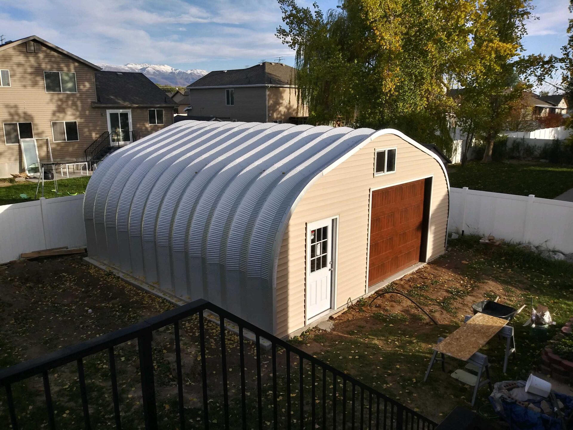 A-Model Quonset hut with tan end wall, brown garage door, white man door, and black railing going down in the front of the photo.