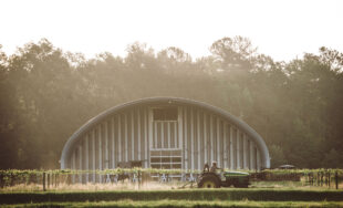 S model steel arch building with steel recessed endwalls used for storage on a farm with guy riding a green tractor in front with morning dew visable
