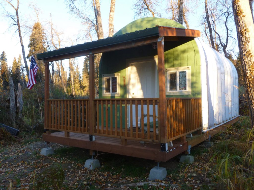 Tiny Quonset hut on elevated wooden platform in woods, custom green endwall, white door, covered front porch with bench and American flag