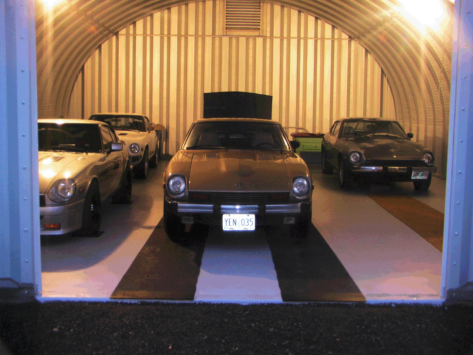 S-Model Quonset hut with four muscle cars inside, two on the right are black, and the two on the left are a lighter color.