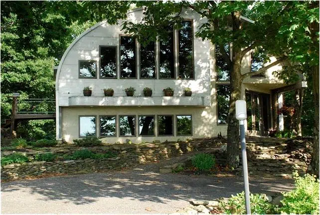 Eco-friendly S-model Quonset home in the woods with a custom tan endwall, large windows, a ledge with potted plants, and a side balcony.