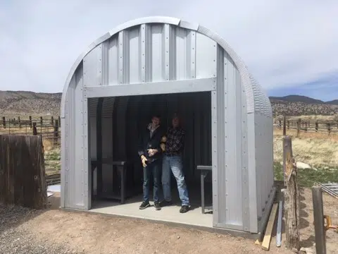 Open blacksmith workshop with steel endwall, two people inside, open land background with wooden fence.