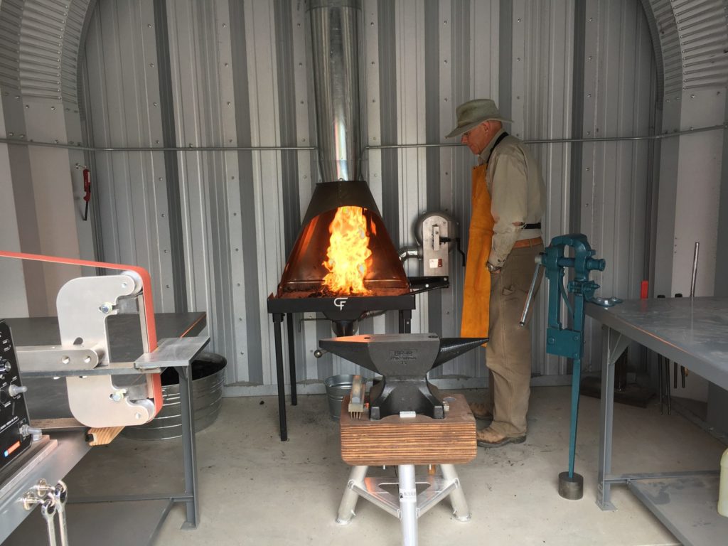 Inside a Quonset workshop with a steel endwall: a blacksmith working over open flame and various blacksmith equipment and tools including an anvil.