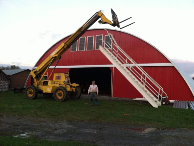 Custom red endwall with upper windows on Q-model Quonset hangar with large center opening, heavy machinery lifting white staircase in front of building.