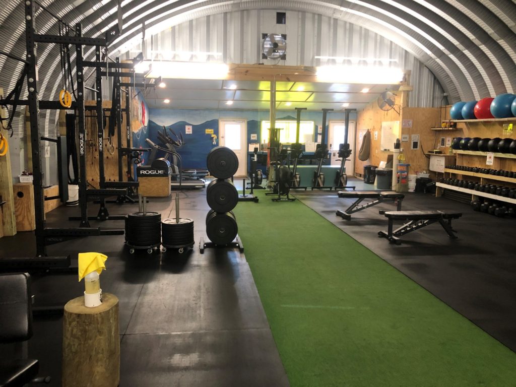 Inside a Quonset hut: partially wooden walls with shelving, padded flooring, various gym equipment inside.