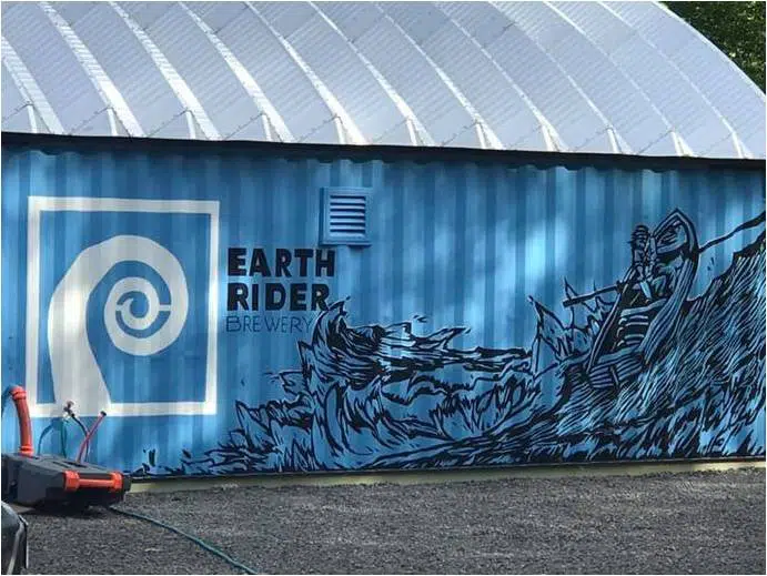 Side of container cover building, metal arch roof system sitting on a blue freight container stamped with Earth Rider Brewery logo and a painting of a person on a rowboat in waves.