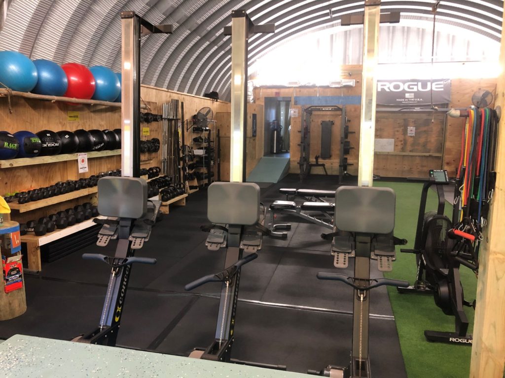 Inside a Quonset hut: partially wooden walls with shelving, padded flooring, various gym equipment inside.