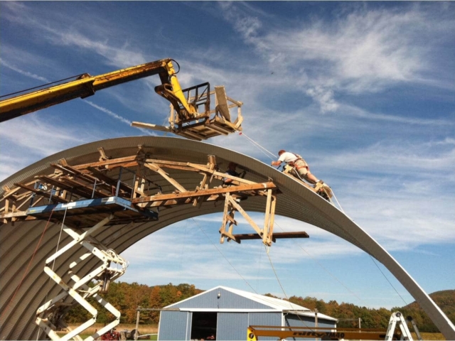 Assembling of large Quonset hangar, lift and wooden trusses holding arches in position while a man works to secure the panel from the top of the structure.