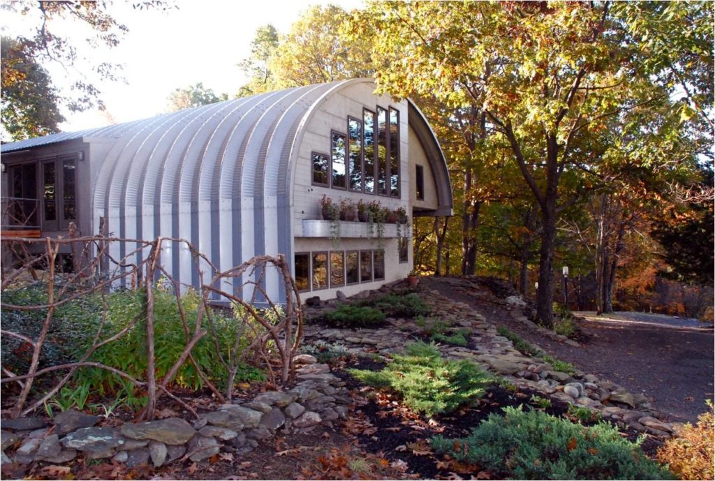 Eco-friendly S-model Quonset home in the woods with a custom tan endwall, large windows, a ledge with potted plants, and a side balcony with entrance and windows.