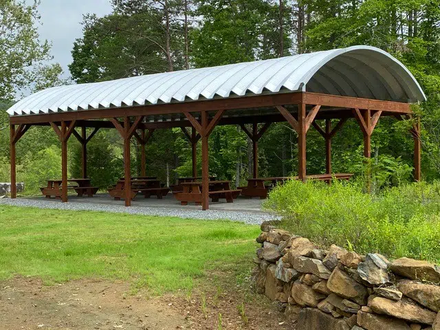 Wooden picnic tables on gravel underneath a Quonset picnic pavilion held up by wooden beams, surrounded by forest.