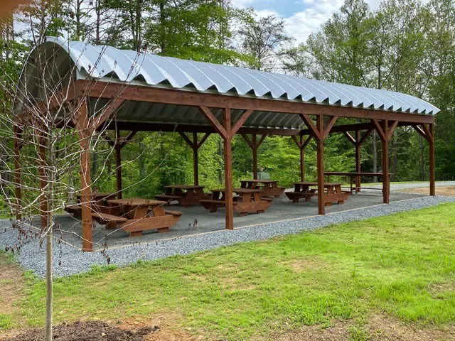 Wooden picnic tables on gravel underneath a Quonset picnic pavilion held up by wooden beams, surrounded by forest.