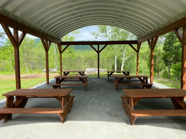 Wooden picnic tables on gravel underneath a Quonset picnic pavilion held up by wooden beams, surrounded by forest.