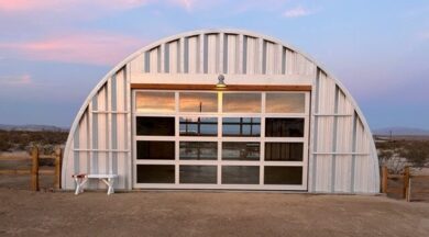Q-Model Quonset hut with steel end walls, glass garage door, a light above the door, and a white bench on the left of the building.