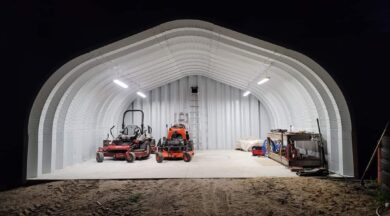 One-ended A-Model Quonset hut with a red and orange lawn mower, a worktable on the right, and ladder leaning against the back.