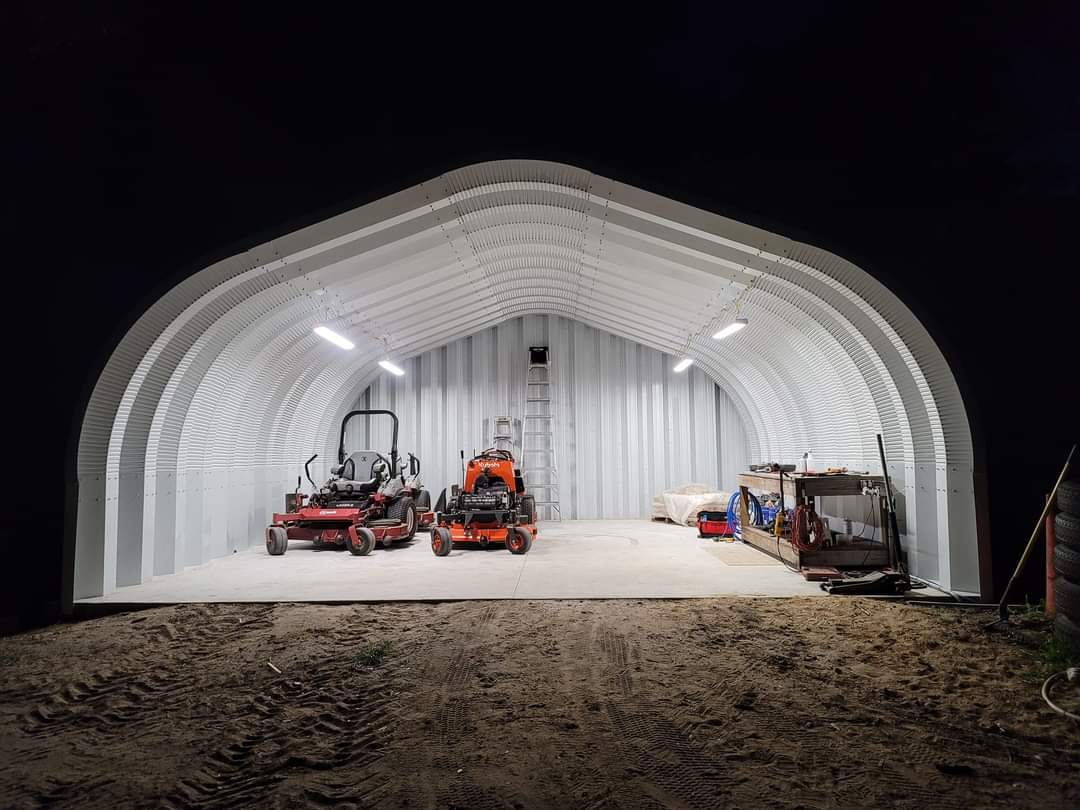 One-ended A-Model Quonset hut with a red and orange lawn mower, a worktable on the right, and ladder leaning against the back.