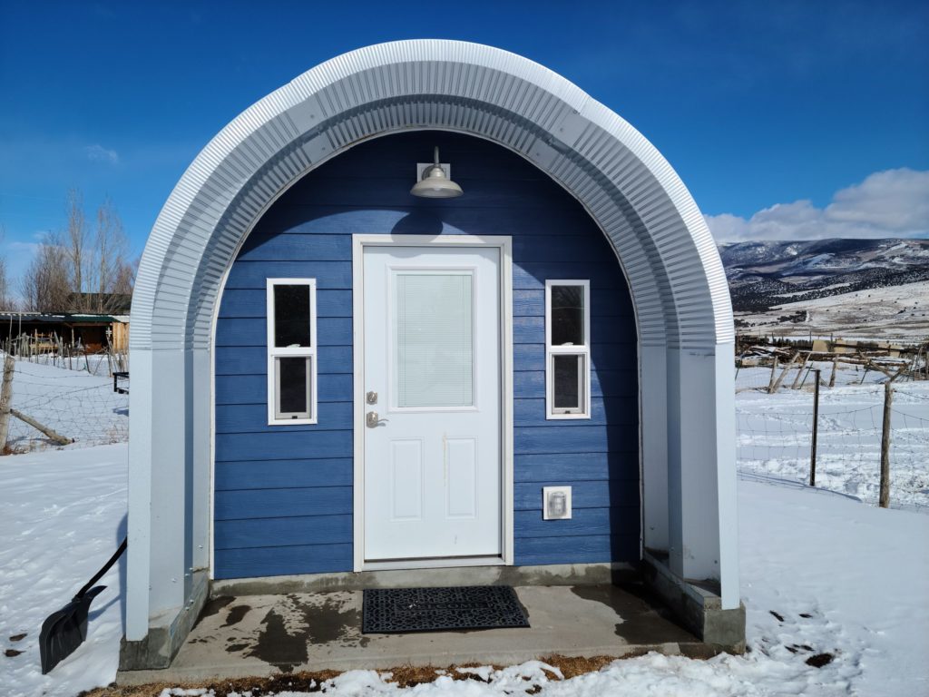 Tiny blue Quonset shed with custom endwall, white door, hanging entrance light, awning over porch, snowy lanscape.