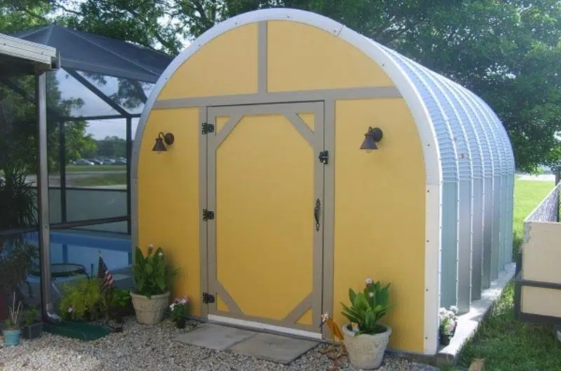 Tiny Quonset hut with custom yellow endwall, entrance door with tan trimming, potted plants at front.
