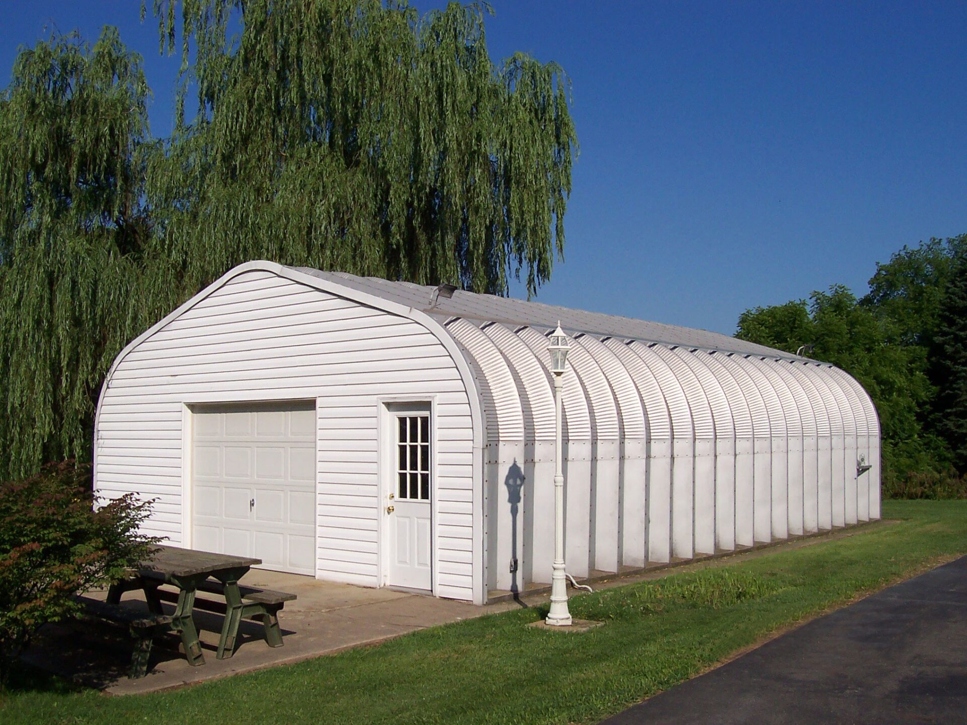 A-Model Quonset hut with white end wall, white garage door, and white lamp pole with a brown bench outside of the Quonset hut.