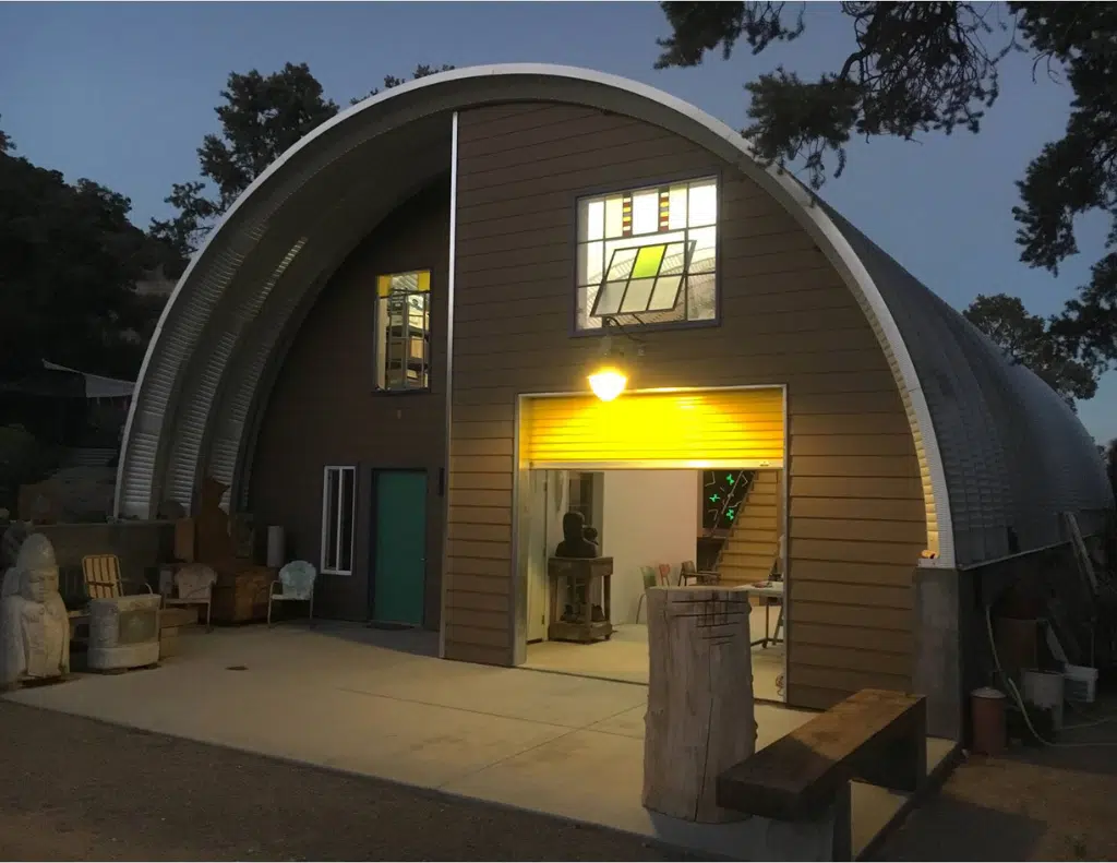 Q-model Quonset home with brown custom endwall partially recessed, overhead awning, arched roof set on raised concrete sidewall, rolling door, green entrance door, upper windows.