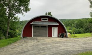 Q-Model Quonset hut with custom red end wall. grey garage door, white man door, and extending roof above the doors, and a window above the extension.