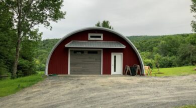 Q-Model Quonset hut with custom red end wall. grey garage door, white man door, and extending roof above the doors, and a window above the extension.