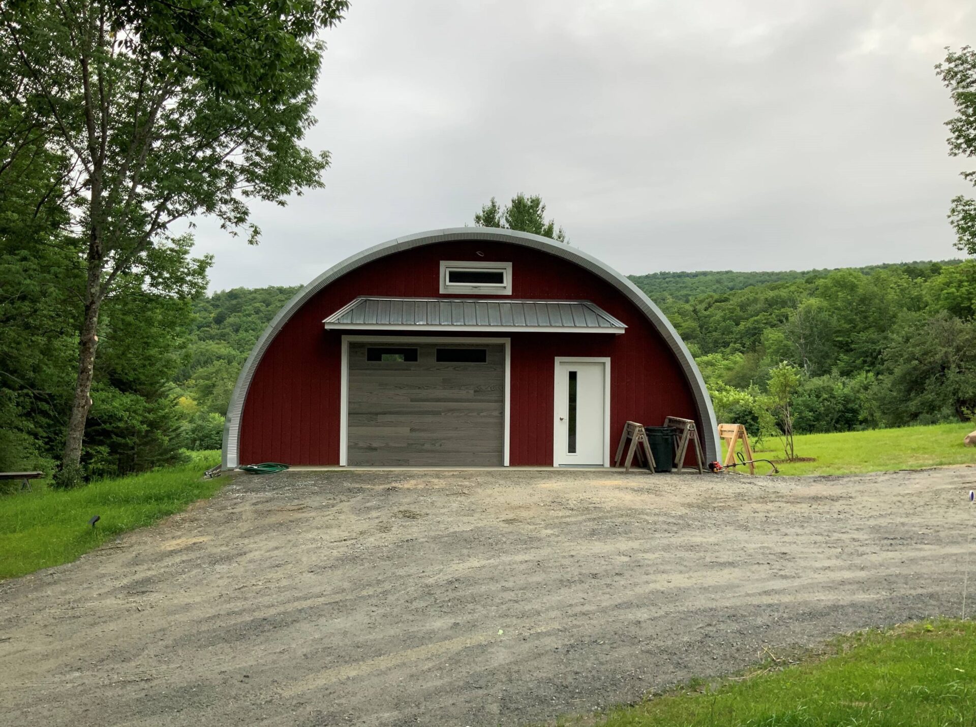 Q-Model Quonset hut with custom red end wall. grey garage door, white man door, and extending roof above the doors, and a window above the extension.