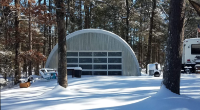 Q-Model Quonset hut with custom made end wall, glass garage door, covered in snow, and an RV parked on the right.