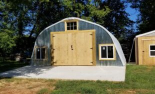 X-Model Quonset hut with custo wooden garage door, windows to the left, right, and top of the garage door, and a wooden shed to the right of the Quonset.