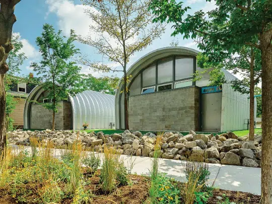 Two Quonset huts with custom endwalls, concrete path, rocks, and trees in front.