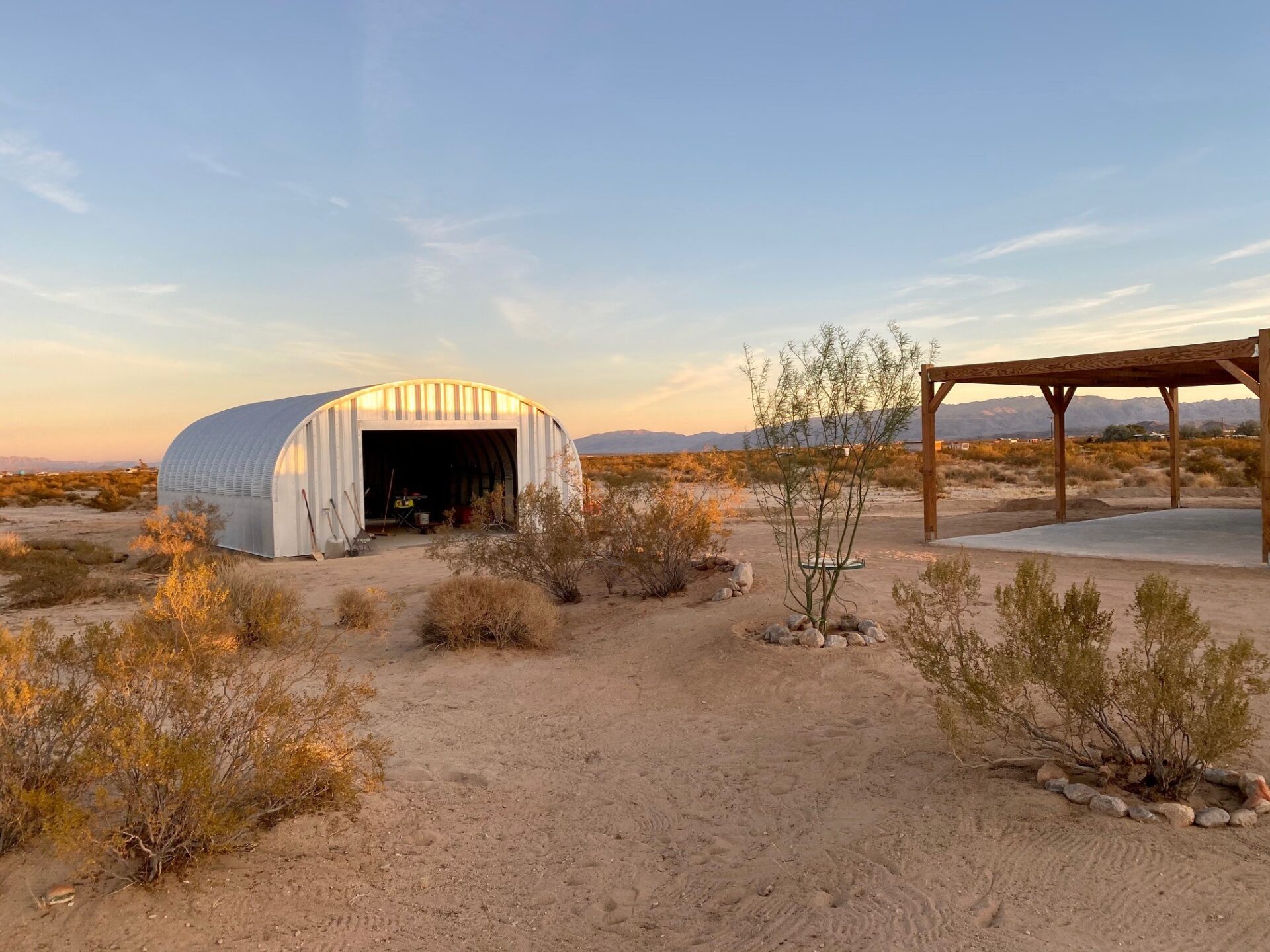S-Model Quonset hut with open garage door, tumbleweeds on the ground, a canopy on the right, and shovels leaning against the building.
