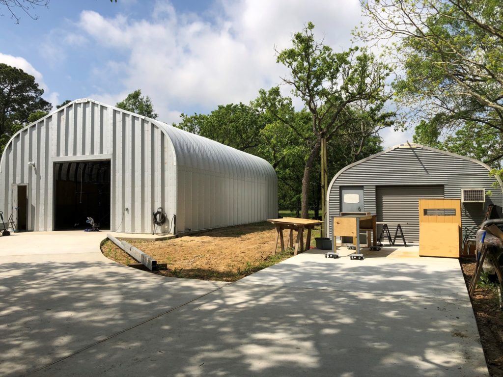 Driveways leading to two A-model Quonset huts: one with steel endwalls, large rolling door, front entrance; the other with custom ribbed grey endwall, matching rolling door, black entrance door, and workshop equipment in front.
