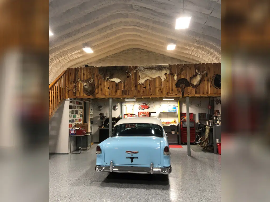 Inside of Quonset workshop: hanging ceiling lights, blue vintage car, wooden loft with hanging animal mounts and skins, antlers, tools and shelving lining walls.