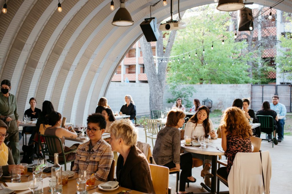 Event space under open-ended Quonset hut, hanging lights and projector, people gathered at tables enjoying food and drinks.