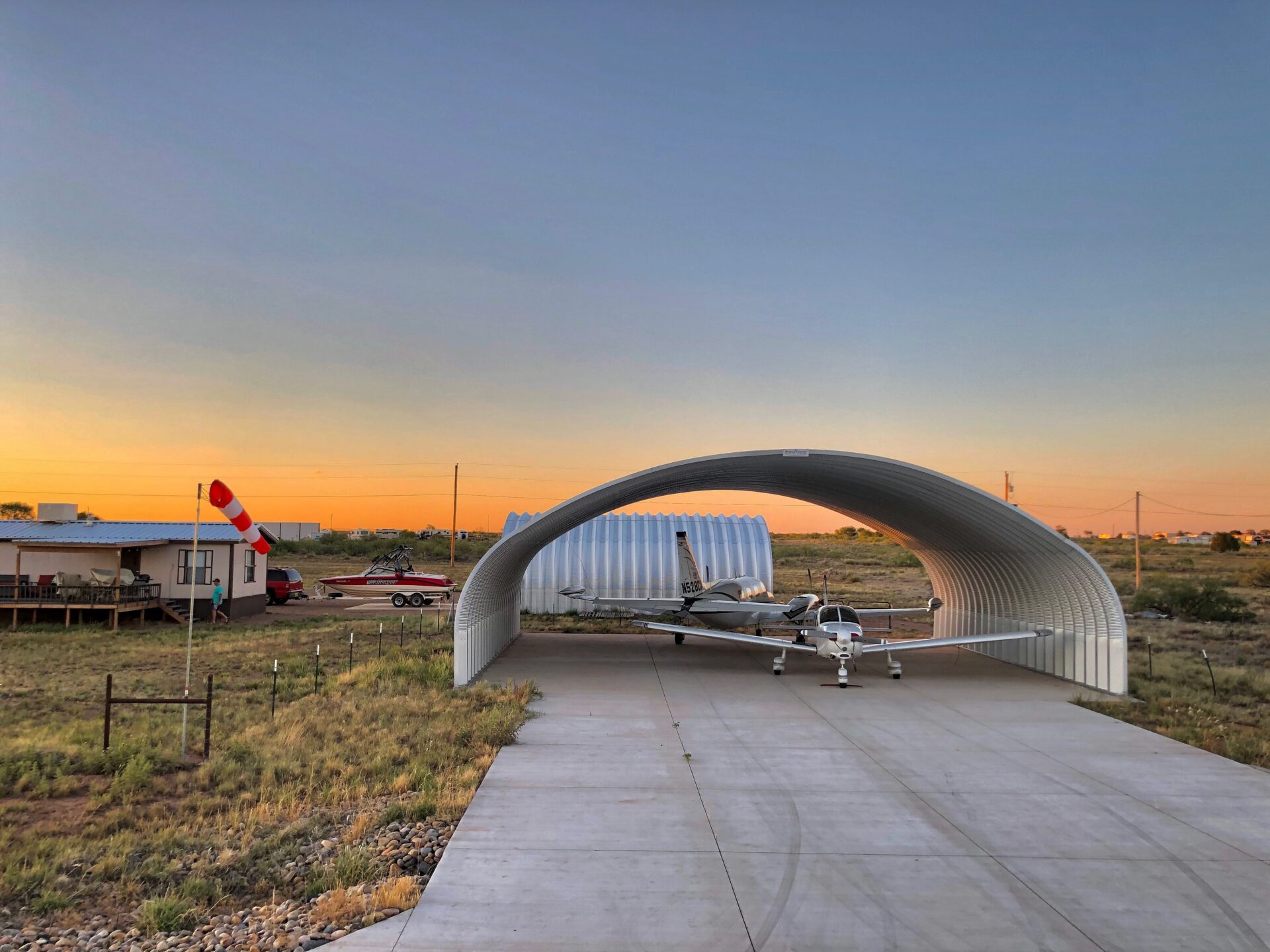 metal quonset hut hangar with open ends and two airplanes inside. another quonset hut can be seen in the background next to a home and red speedboat