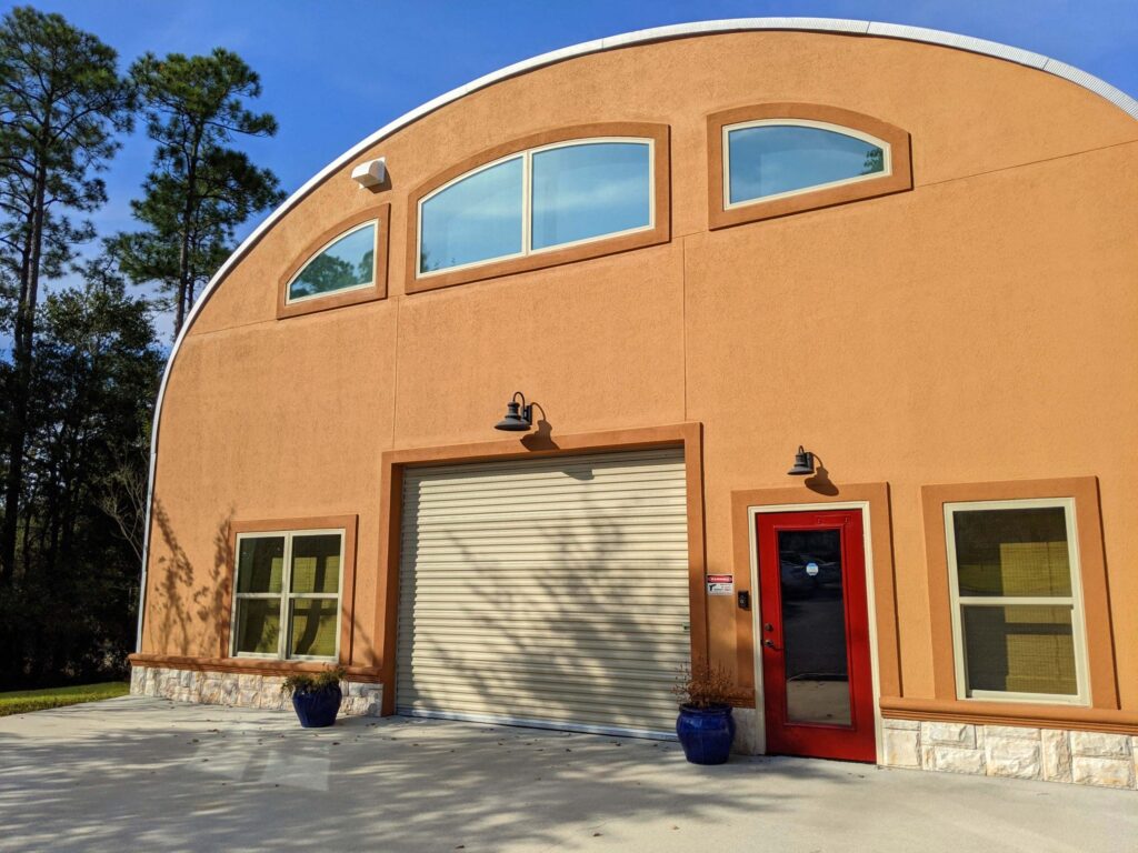 Orange S-model Quonset home, red front door, rolling door, front lower and upper windows, potted plants.