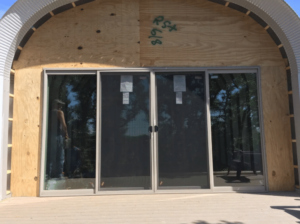 Front of a Quonset casita under construction with custom wooden endwall, glass french doors, and two large windows to either side.