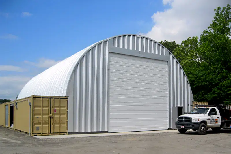Metal container cover workshop set on top of freight containers, steel endwall, front entrance, large garage door with white truck out front.