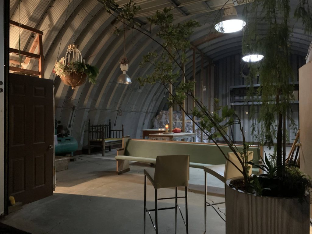 Inside Quonset workshop: hanging overhead lights, potted plants, benches, other stored items.