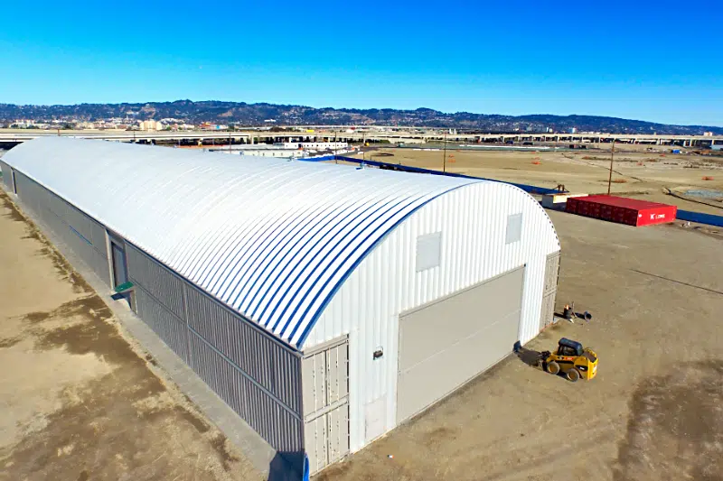 Steel commercial container project: steel roof arches, tan shipping containers, tan garage door, white metal endwall with yellow construction equipment near the garage door.