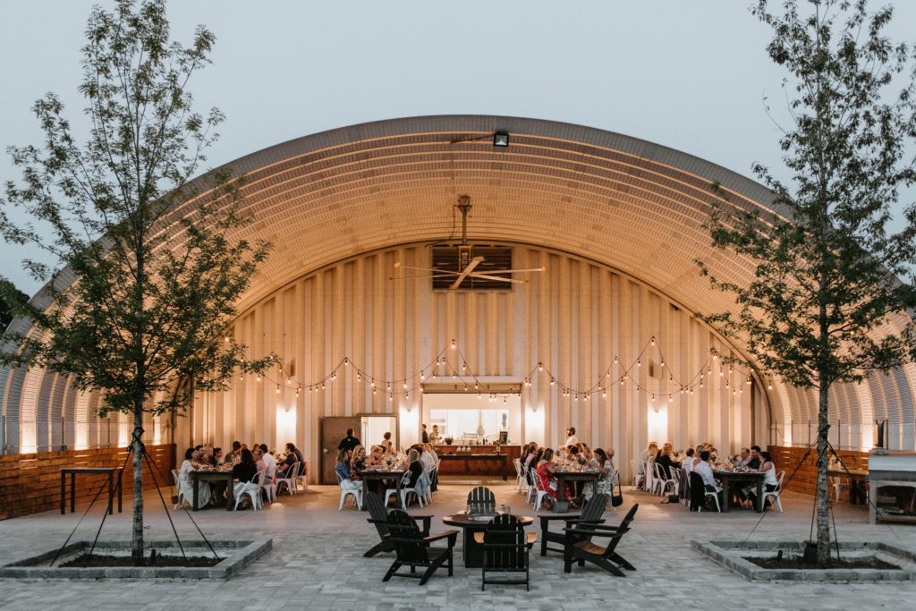 Open ended S-model food pavilion, large ceiling fan, suspended lights, tables filled with people dinning in.