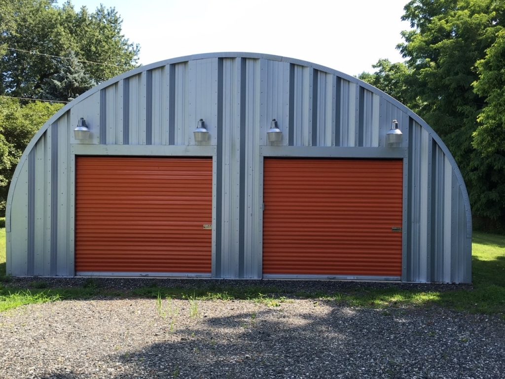 Quonset garage with steel endwall and two copper colored rolling doors, light fixtures above entryways, gravel driveway