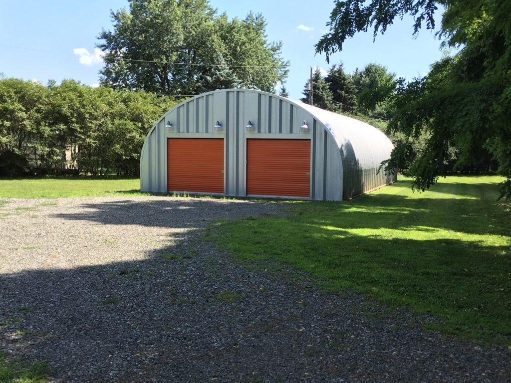 Quonset garage with steel endwall and two copper colored rolling doors, light fixtures above doors, gravel driveway
