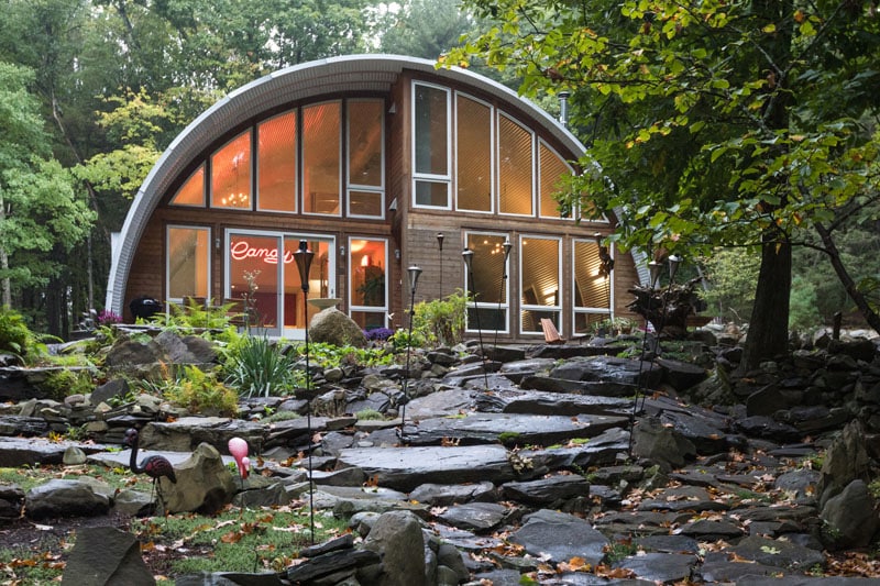 Q-model Quonset home in woods with custom endwall made of wood and large windows, rock path leading up to house.