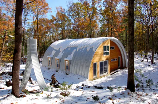 Q-model Quonset home in a forest with snow covered ground, three windows in the side metal arches, custom endwall of wood, windows, and sliding glass doors. Two arch pieces vertically lean together near a tree in front.