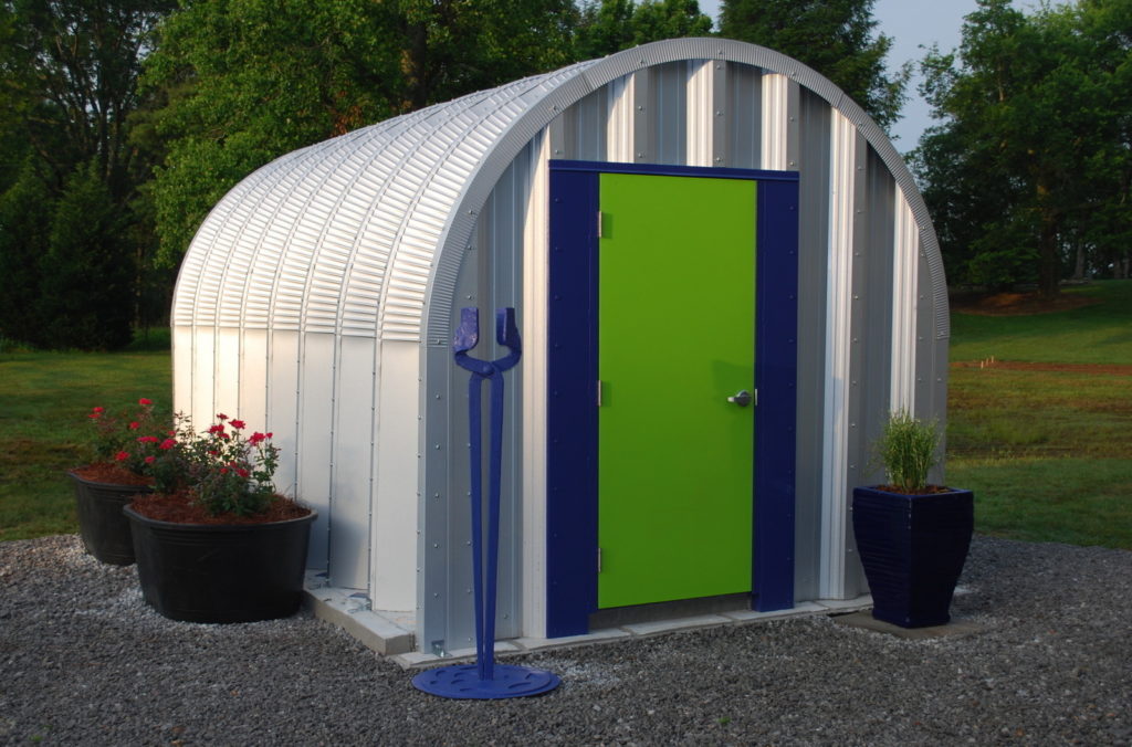 Tiny S-model Quonset hut with steel endwall, green door trimmed in navy, potted plants in front and to side of building.