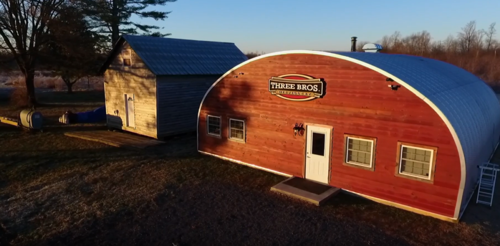 S-model Quonset hut distillery with a custom red wood endwall, white entrance door, and windows across the front.